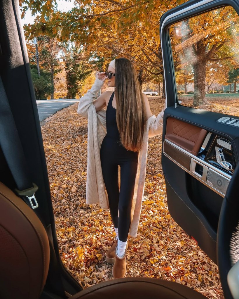 Woman in black bodysuit and cardigan standing on autumn leaves from a car.