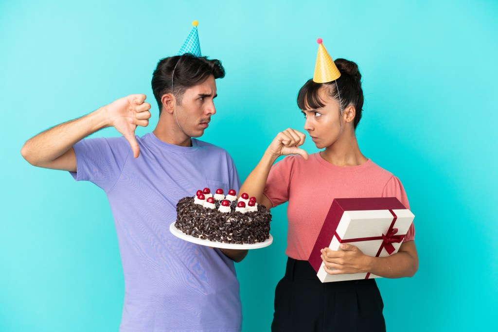A young couple dressed in holiday hats gives a finger down while wearing a birthday cake and a gift.
